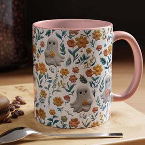 May include: White ceramic mug with a pink interior and handle. The mug features a repeating pattern of cartoon ghosts holding flowers, interspersed with orange and yellow flowers and green leaves. The mug is sitting on a wooden surface.
