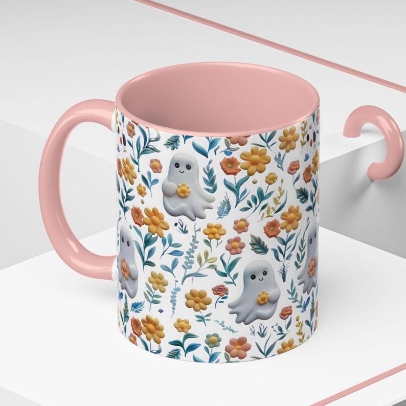 May include: A ceramic mug with a pink handle and interior. The exterior features a pattern of smiling ghost figures holding flowers, interspersed with orange, yellow, and pink flowers, and green leaves on a white background. A whimsical design.