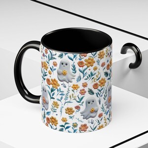 May include: A ceramic mug with a black handle and interior. The exterior features a repeating pattern of cartoon ghosts holding flowers, set against a backdrop of orange and yellow flowers and green leaves on a white background.