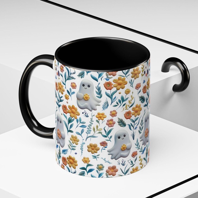 May include: A ceramic mug with a black handle and interior. The exterior features a repeating pattern of cartoon ghosts holding flowers, set against a backdrop of orange and yellow flowers and green leaves on a white background.