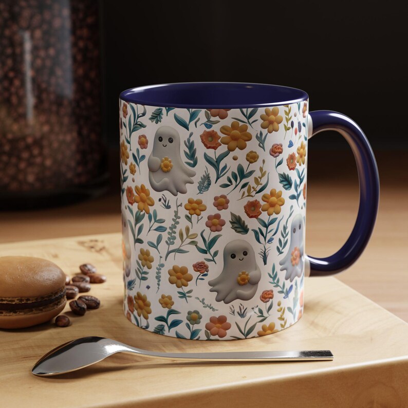 May include: A ceramic mug with a blue handle and interior. The exterior features a repeating pattern of gray cartoon ghosts holding yellow flowers, interspersed with orange and yellow flowers and green leaves on a white background.