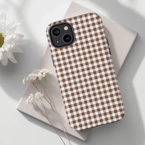 Può includere: Custodia per telefono con motivo a quadretti marrone e bianco. La custodia è su una superficie grigio chiaro con fiori bianchi sullo sfondo. La custodia del telefono ha un modulo fotocamera nero.