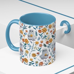May include: A ceramic mug with a light blue interior and handle. The exterior features a white background with a floral pattern of orange and yellow flowers, green leaves, and smiling ghost illustrations. The mug is designed for beverages.