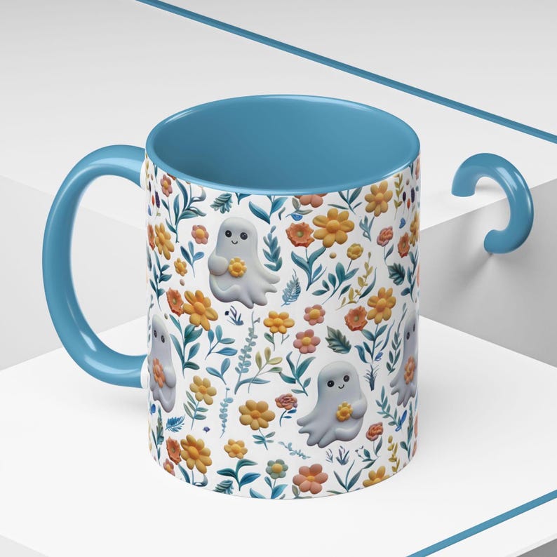 May include: A ceramic mug with a light blue interior and handle. The exterior features a white background with a floral pattern of orange and yellow flowers, green leaves, and smiling ghost illustrations. The mug is designed for beverages.