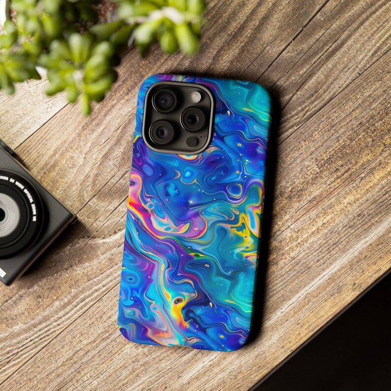 May include: A smartphone case with a vibrant, abstract design. The case features a swirling pattern of blues, purples, yellows, and pinks, creating a colorful, marbled effect. The case has a camera cutout and is set on a wooden surface.