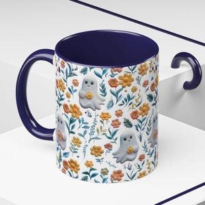 May include: A ceramic mug with a navy blue interior and handle. The exterior features a white background with a repeating pattern of orange and yellow flowers, green leaves, and smiling ghost illustrations. The mug is designed for hot beverages.