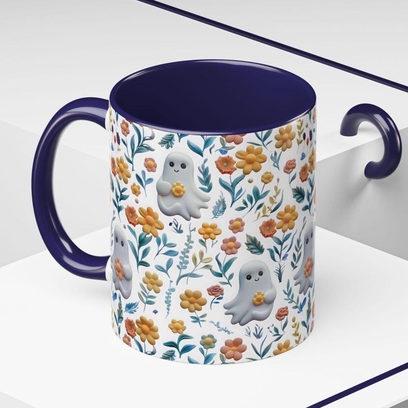 May include: A ceramic mug with a navy blue interior and handle. The exterior features a white background with a repeating pattern of orange and yellow flowers, green leaves, and smiling ghost illustrations. The mug is designed for hot beverages.