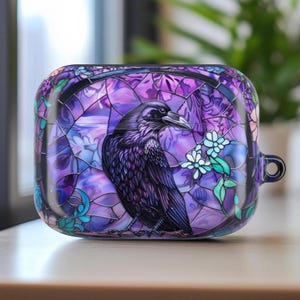 May include: A purple and black protective case featuring a raven design. The case has a stained-glass style with a raven perched on a branch with white and green flowers. The case is rectangular with rounded edges and a small loop.