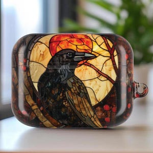 May include: A decorative case for wireless earbuds featuring a stained-glass design. The artwork depicts a black raven perched against a backdrop of a setting sun and bare tree branches, with red berries. The case has a glossy finish.