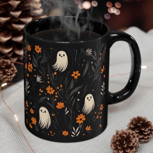 May include: Black ceramic mug featuring a whimsical design of smiling ghosts, orange flowers, and foliage. The mug has a black handle and is filled with a hot beverage, with steam rising from the top. A cozy, seasonal theme.