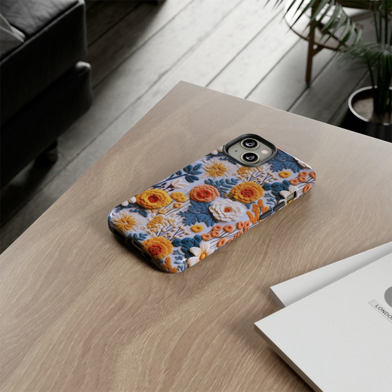 Pode incluir: Uma capa de telefone branca com um padr&atilde;o floral com flores amarelas, laranja e azuis. A capa tem uma protuber&acirc;ncia de c&acirc;mera elevada com duas lentes pretas.