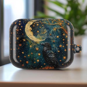May include: A black and navy blue AirPod case featuring a detailed illustration of a raven perched beneath a crescent moon and stars. The design includes a tree and a starry night sky, with gold accents.