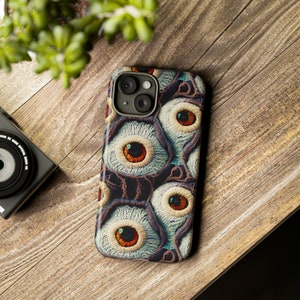 May include: A phone case with a colorful, abstract design featuring multiple eyes. The case is made of a durable material and has a raised camera bump for protection.