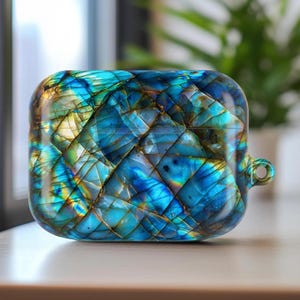 Può includere: Una custodia quadrata per auricolari wireless. La custodia è prevalentemente blu con striature gialle, verdi e dorate. La superficie presenta un motivo geometrico e fratturato e sul lato è visibile un piccolo anello.