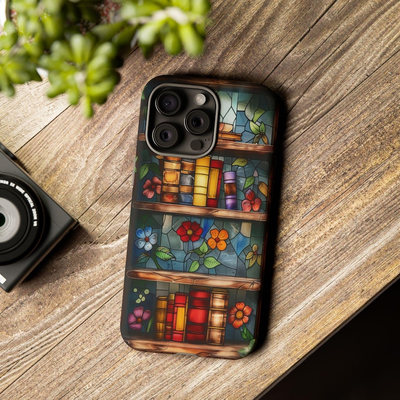 May include: A phone case with a stained glass design featuring a bookshelf with colourful books and flowers. The case has a camera lens cutout and a black border.