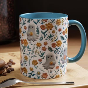 May include: A ceramic mug with a white background, adorned with a floral pattern of orange and yellow flowers, green leaves, and cute ghost illustrations. The interior and handle are a solid teal color.