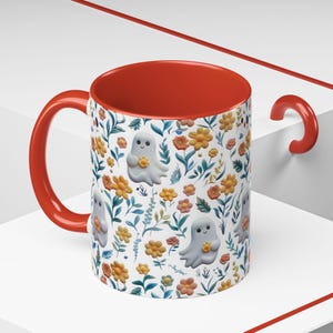 May include: A ceramic mug with a red interior and handle. The exterior features a pattern of cartoon ghosts holding flowers, interspersed with orange and yellow flowers and green leaves on a white background. The mug is designed for beverages.
