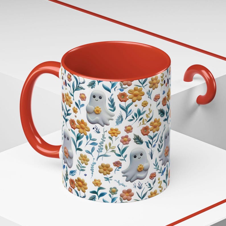 May include: A ceramic mug with a red interior and handle. The exterior features a pattern of cartoon ghosts holding flowers, interspersed with orange and yellow flowers and green leaves on a white background. The mug is designed for beverages.
