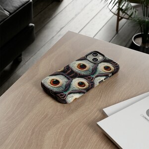May include: A black phone case with a pattern of multiple eyes in shades of brown, white, and blue. The case is designed for an iPhone.