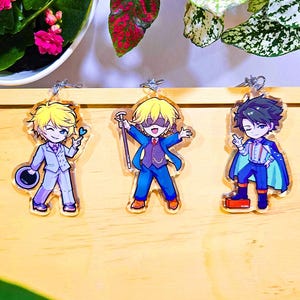 Op de afbeelding: Drie acryl sleutelhangers in anime-stijl met mannelijke personages in verschillende outfits. De personages hebben blond en donker haar en dragen pakken, capes en houden accessoires vast. De sleutelhangers liggen op een houten oppervlak met planten op de achtergrond.