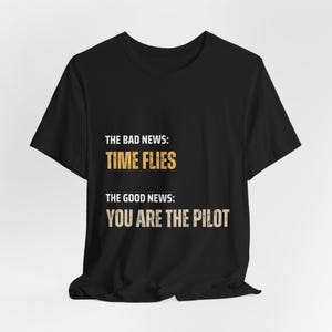 Puede incluir: Camiseta negra con el texto "THE BAD NEWS: TIME FLIES" y "THE GOOD NEWS: YOU ARE THE PILOT" en amarillo.
