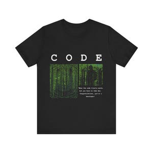 Op de afbeelding: Zwart T-shirt met een groene en zwarte afbeelding van code en de tekst "CODE" boven de afbeelding. De tekst onder de afbeelding luidt: "Wanneer de code eindelijk werkt, maar je hebt geen idee waarom. Gefeliciteerd, je bent een ontwikkelaar!"