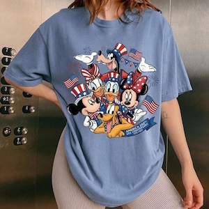 A woman wearing a blue t-shirt with a graphic of Disney characters, including Mickey Mouse, Minnie Mouse, Donald Duck, Goofy, and Pluto, along with American flags and fireworks.
