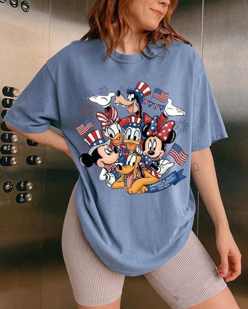 A woman wearing a blue t-shirt with a graphic of Disney characters, including Mickey Mouse, Minnie Mouse, Donald Duck, Goofy, and Pluto, along with American flags and fireworks.