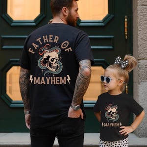 May include: Matching black t-shirts with a skull and snake graphic. The adult shirt reads "FATHER OF MAYHEM" on the back. The child's shirt has the same design with "MAYHEM" below. Both shirts are black.