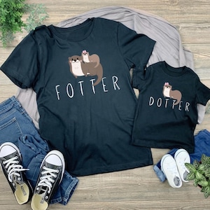 May include: Two black t-shirts with otter graphics. One shirt says "FOTTER" and has a larger otter graphic with two otters. The other shirt says "DOTTER" and has a smaller otter graphic. The shirts are on a wooden surface.