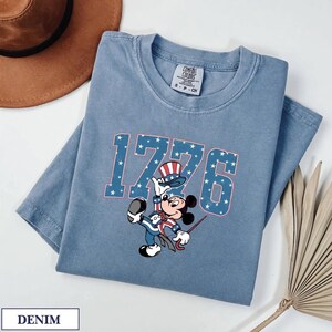 Mickey Mouse in patriotic attire with "1776" in star-patterned letters on a denim t-shirt.