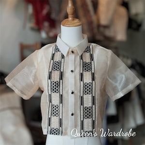 Op de afbeelding: Een crèmekleurig shirt met korte mouwen, een witte kraag en zwarte knopen. Het shirt heeft een gedetailleerd, geometrisch patroon in zwart en crème op de voorkant. De tekst "Queen's Wardrobe" is onderaan zichtbaar.