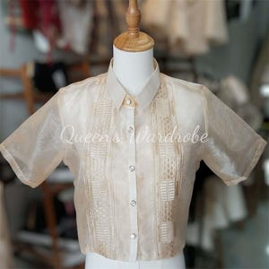 Op de afbeelding: Een beige blouse met korte mouwen en ingewikkelde geborduurde details aan de voorkant en op de mouwen. De blouse heeft een kraag en is aan de voorkant dichtgeknoopt met doorzichtige knopen. De tekst "Queen's Wardrobe" is zichtbaar op de voorkant.