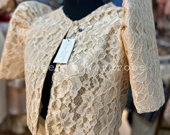 May include: A cream-coloured lace bolero jacket with short, puffed sleeves. The jacket features a round neckline and a single button fastening. The text "Queen's Wardrobe" is visible. The lace has an intricate floral design.
