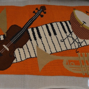 Könnte beinhalten: Kreuzstich-Kunstwerk mit Musikinstrumenten auf orangefarbenem Hintergrund. Eine braune Geige, eine Klaviatur, eine Trommel, eine Trompete und ein Waldhorn sind dargestellt. Das Kunstwerk ist mit "Shari '75" signiert.
