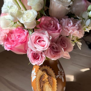 Op de afbeelding: Een boeket van roze en witte rozen, anjers en andere bloemen in een bruine vaas. De vaas heeft een gesneden houten afbeelding van de Maagd Maria en witte madeliefachtige ontwerpen.