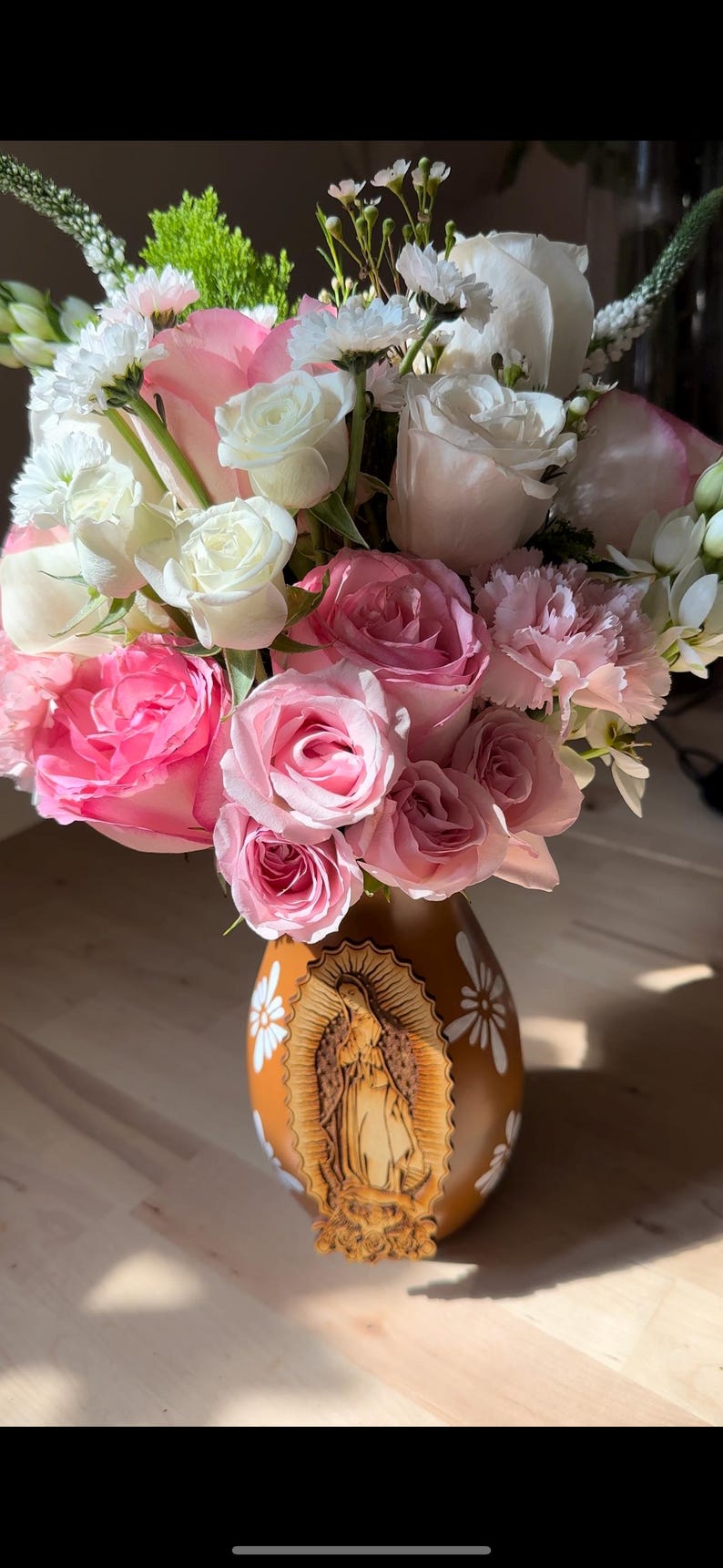 Op de afbeelding: Een boeket van roze en witte rozen, anjers en andere bloemen in een bruine vaas. De vaas heeft een gesneden houten afbeelding van de Maagd Maria en witte madeliefachtige ontwerpen.