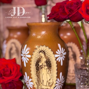 Op de afbeelding: Een bruine keramische vaas met witte madeliefachtige ontwerpen en een houten uitsnede van de Maagd Maria. Rode rozen omringen de vaas. De tekst "JD Designs - LASER CUT & ENGRAVE" is zichtbaar.