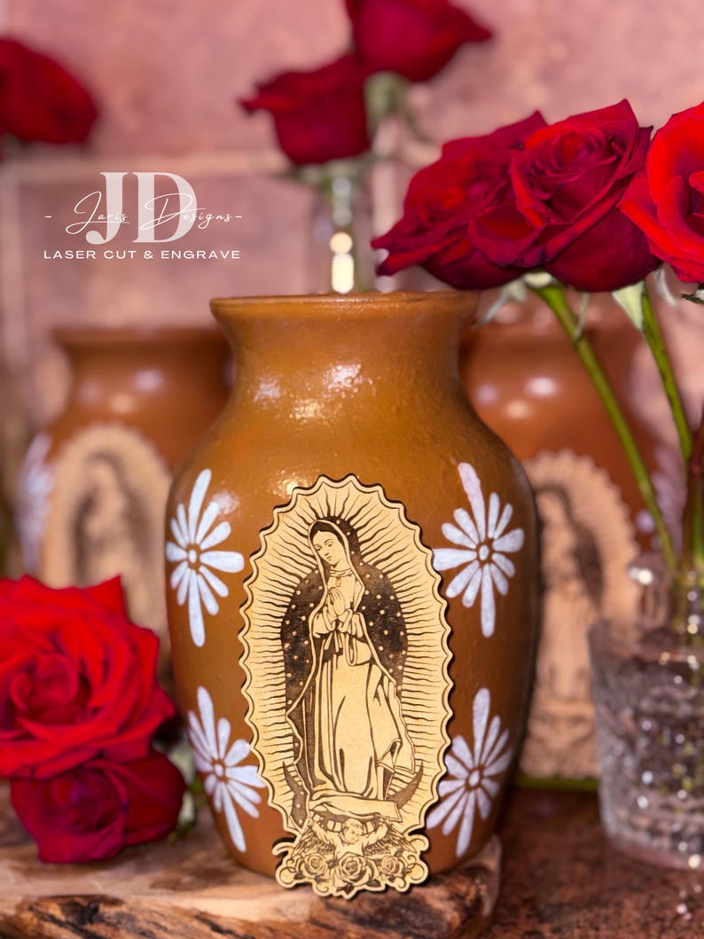 Op de afbeelding: Een bruine keramische vaas met witte madeliefachtige ontwerpen en een houten uitsnede van de Maagd Maria. Rode rozen omringen de vaas. De tekst "JD Designs - LASER CUT & ENGRAVE" is zichtbaar.