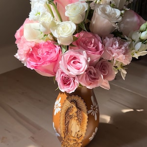 Op de afbeelding: Een bloemstuk met roze en witte rozen, anjers en andere bloemen in een bruine vaas. De vaas heeft een gesneden afbeelding van een religieuze figuur en witte bloemmotieven. De bloemen zijn gerangschikt in een vol, weelderig boeket.