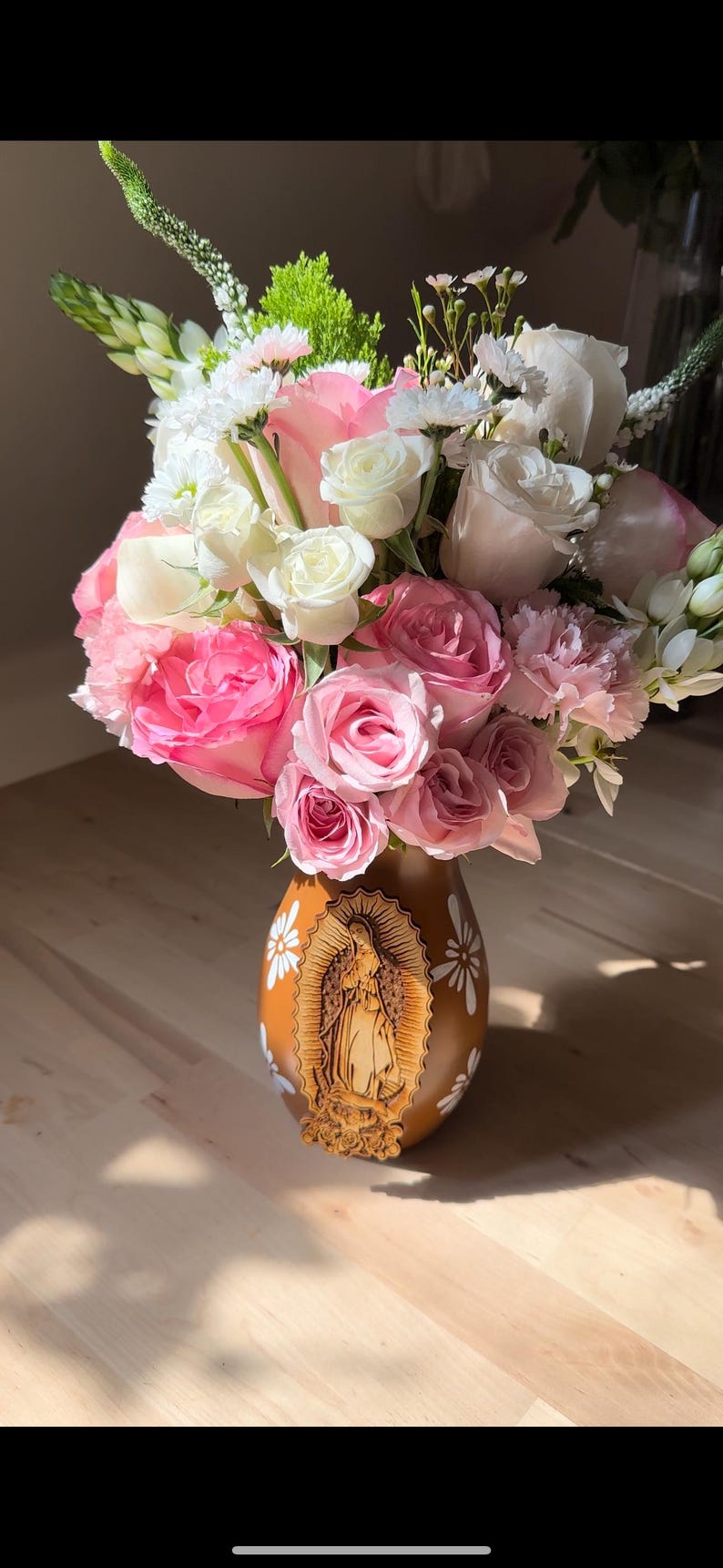 Op de afbeelding: Een bloemstuk met roze en witte rozen, anjers en andere bloemen in een bruine vaas. De vaas heeft een gesneden afbeelding van een religieuze figuur en witte bloemmotieven. De bloemen zijn gerangschikt in een vol, weelderig boeket.