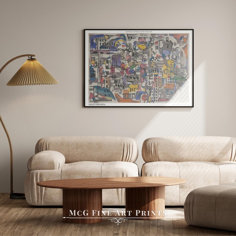 May include: A colorful abstract painting of a city scene with buildings, streets, and vehicles. The painting is framed in black and hangs on a beige wall above a light brown couch and coffee table. The text "The Museum of Modern Art New York 1939" is printed at the top of the painting.
