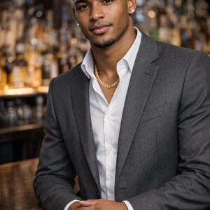 May include: A man in a gray suit, white dress shirt, and gold chain necklace. The man is leaning on a wooden bar. The background is a blurred view of a bar with bottles.