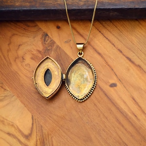 May include: Gold-toned oval locket necklace, open to reveal a dark interior. The locket has a beaded border and hangs from a delicate gold chain. The background is a wooden surface.