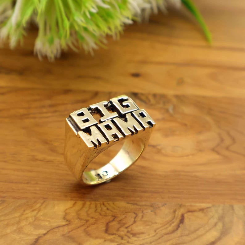 Op de afbeelding: Een goudkleurige ring met de woorden "BIG MAMA" in blokletters. De ring is rechthoekig met een dikke band en ligt op een houten oppervlak. Een opvallend sieraad.