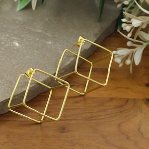May include: A pair of gold-toned stud earrings featuring a three-dimensional wireframe cube design. Each earring is constructed from thin, polished metal rods forming an open geometric square structure with a small post backing.