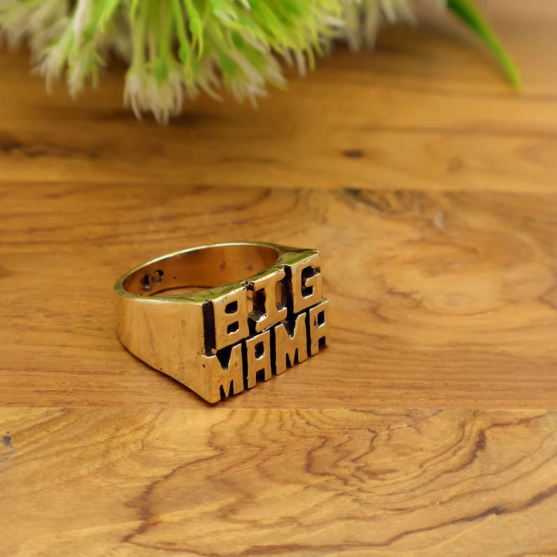 Op de afbeelding: Een goudkleurige ring met de woorden "BIG MAMA" in blokletters. De ring ligt op een houten oppervlak, met een vage groen-witte plant op de achtergrond. Een opvallend sieraad.