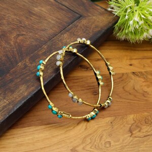 May include: Two gold-colored bangles with turquoise and white gemstone accents. The bangles are resting on a wooden surface. The jewelry is a fashion accessory.
