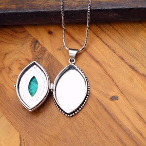 May include: Open silver locket pendant with a turquoise stone and a silver chain. The locket is oval-shaped with a beaded border. The background is a wooden surface.