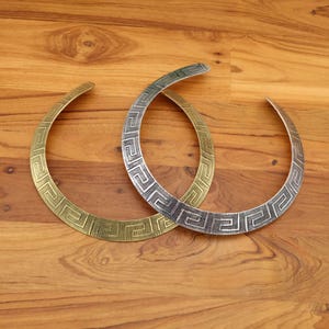 Pode incluir: Dois colares de metal com um padrão de chave grega. Um é dourado, o outro prateado. Os colares são exibidos em uma superfície de madeira, mostrando seu design aberto e detalhes intrincados.